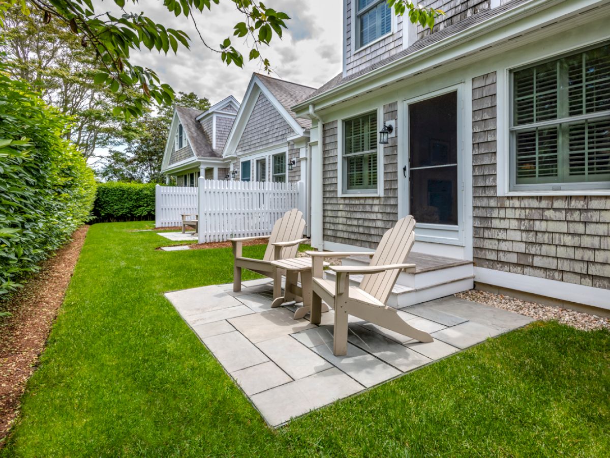 A backyard with two wooden chairs on a stone patio, lush green grass, and a hedge lining the left side of the property, adjacent to a cozy house.