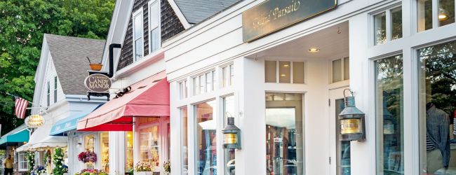 A quaint street lined with charming shops, each featuring bright awnings and inviting storefronts. Colorful flowers in pots adorn the sidewalk.