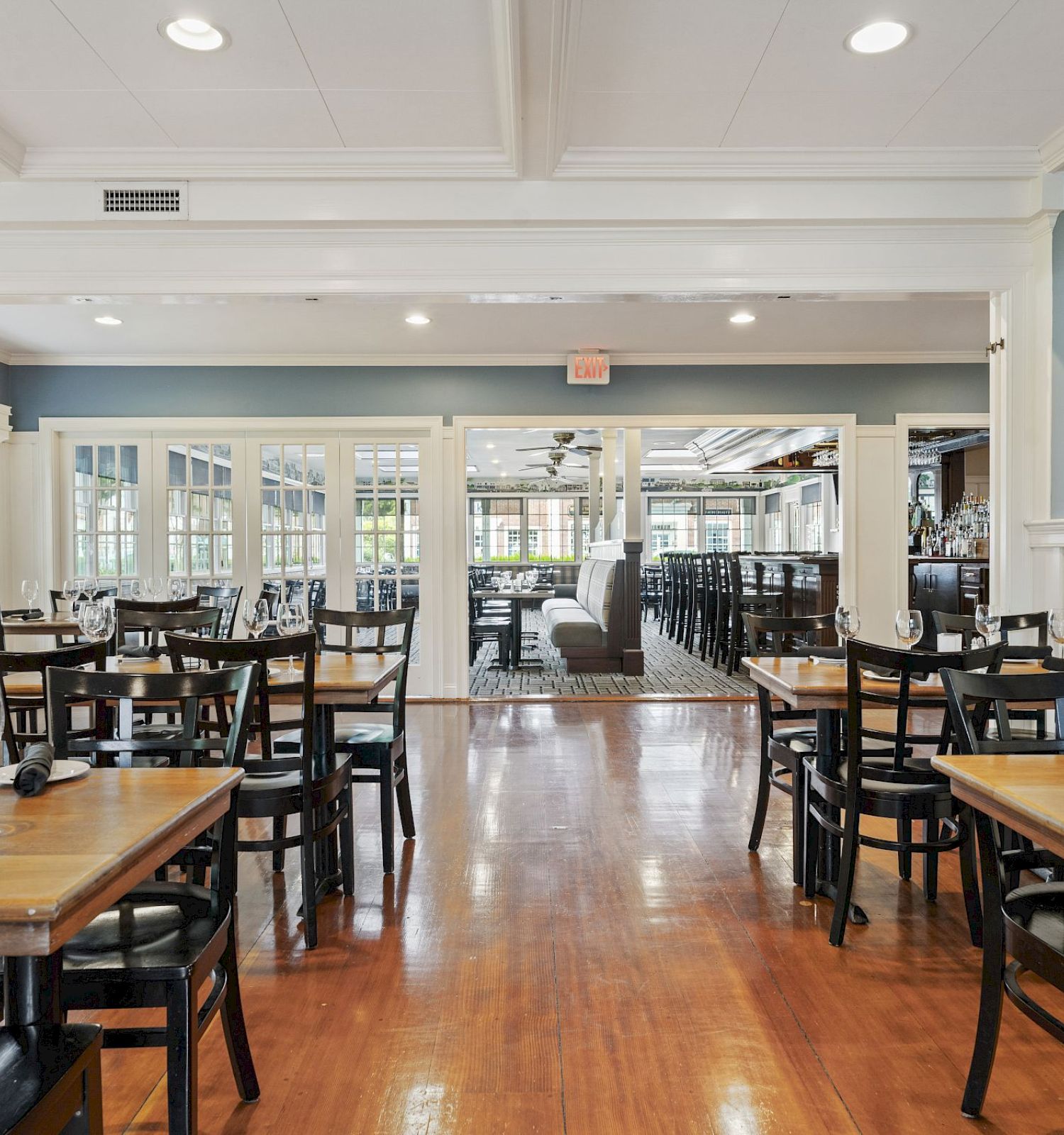 The image shows a spacious, neatly arranged restaurant dining area with wooden floors, empty tables, glassware, and a bright, airy atmosphere.