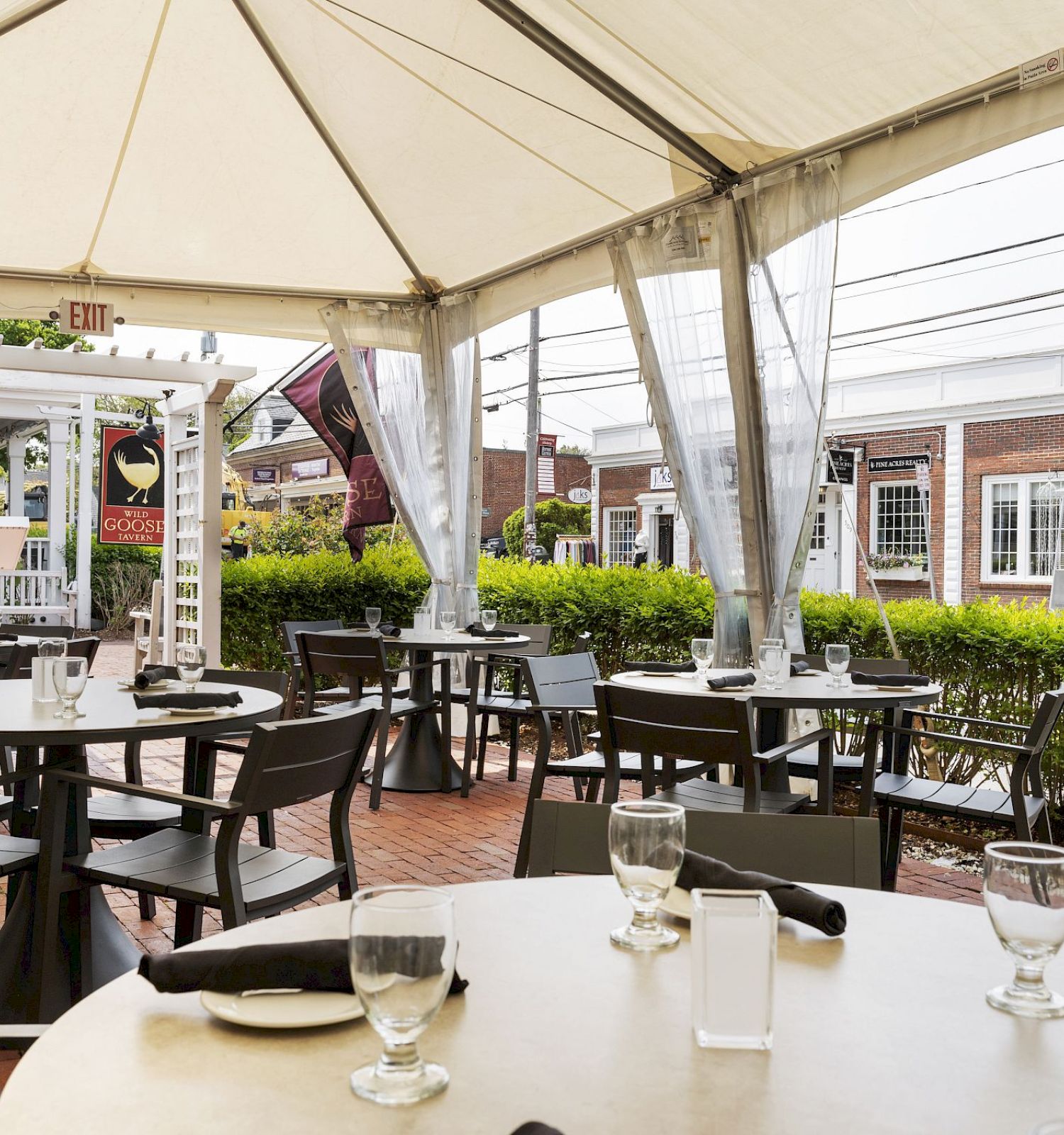 The image shows an outdoor dining area with several round tables covered by a tent, featuring black chairs, glassware, and white napkins.