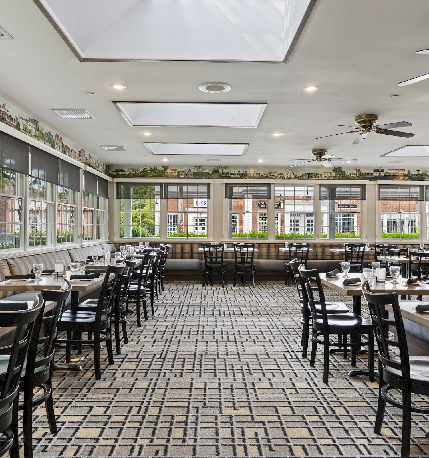 A spacious restaurant dining area with skylights, large windows, and neatly arranged tables and chairs, each set with glasses and napkins.