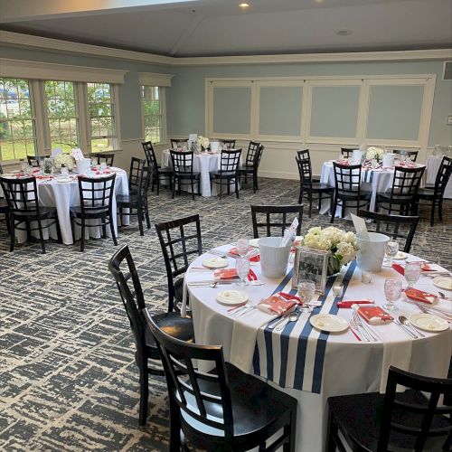 A banquet hall with round tables covered in white tablecloths, decorated with flowers and tableware, set for an event or gathering, is shown.