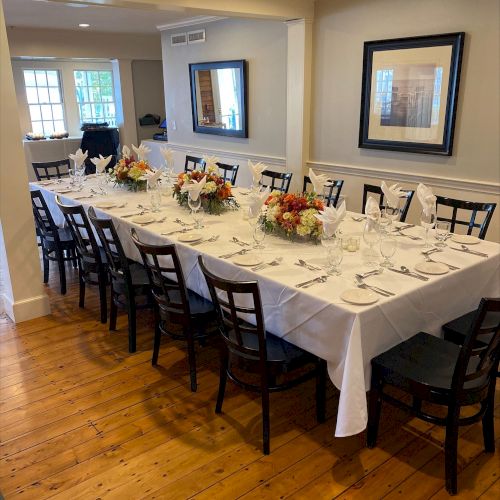 A dining room with a long table set for a formal meal, adorned with flowers and surrounded by black chairs, in a well-lit space.