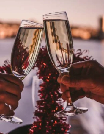 Two hands are toasting with champagne glasses in a festive setting, with blurred cityscape in the background at sunset.