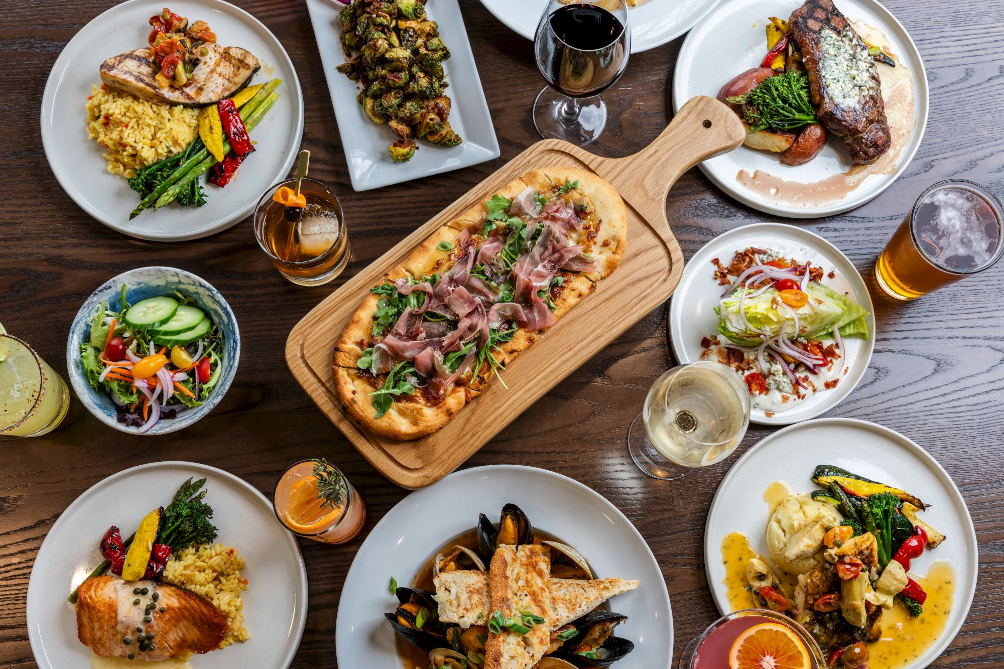 The image shows a table set with various dishes, including grilled meats, salads, vegetables, drinks, and a flatbread, all beautifully arranged.