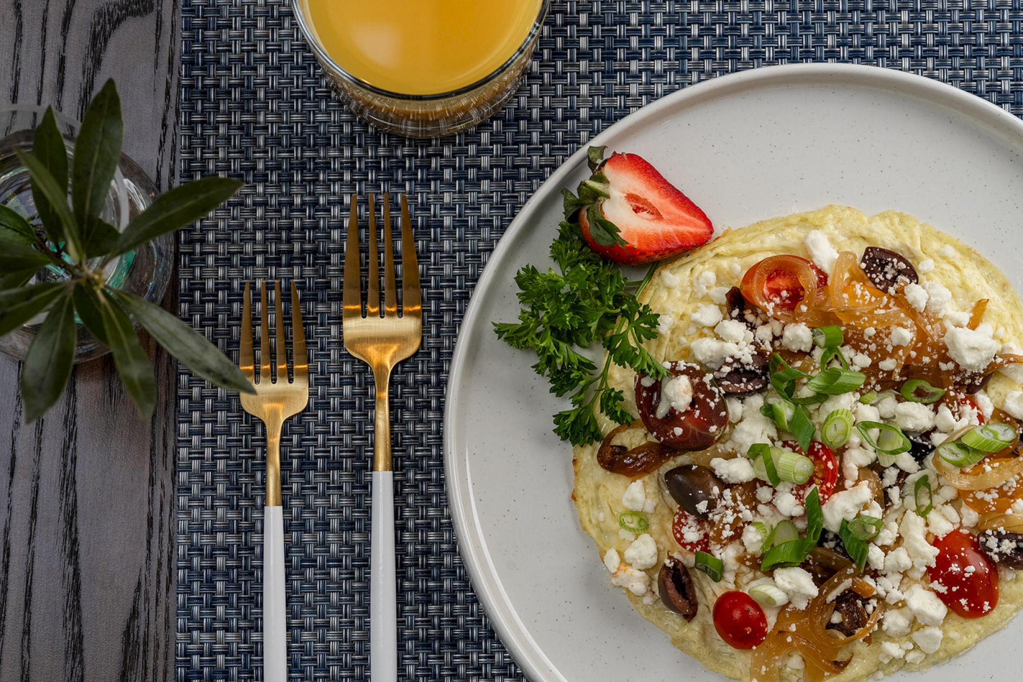 A plated meal with vegetables and cheese, garnished with herbs and strawberries, with a glass of juice, two gold forks, and a plant nearby.