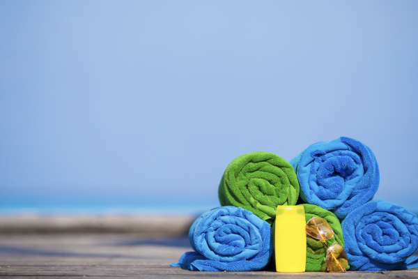 Rolled green and blue towels, yellow bottle, and seashells are on a wooden surface with a blurred sea and sky background.