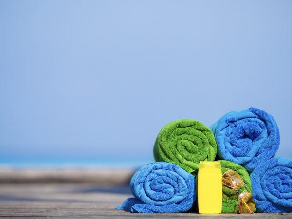 Rolled green and blue towels, yellow bottle, and seashells are on a wooden surface with a blurred sea and sky background.