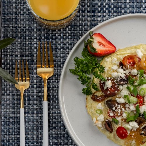 A plate with a colorful omelette topped with vegetables and cheese, garnished with parsley and strawberries, beside gold cutlery, a plant, and a glass of juice.