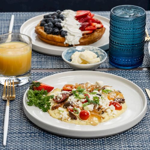 The image shows a breakfast spread with eggs, vegetables, waffles topped with berries, orange juice, a muffin, and a glass of water ending the sentence.