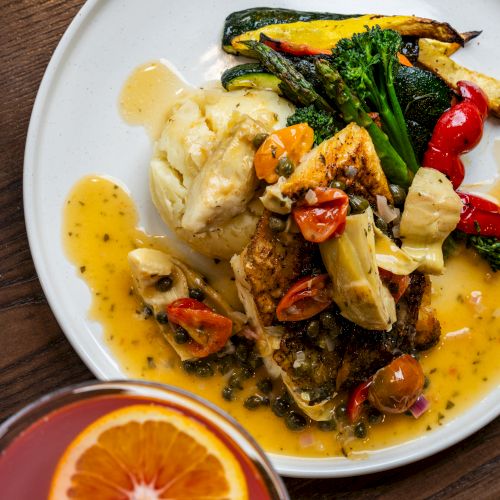 A plate of grilled fish with vegetables and mashed potatoes, covered in a yellow sauce, and a glass of cocktail with an orange slice.