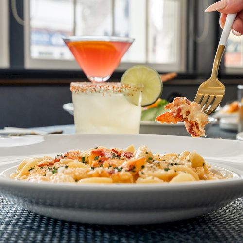 The image shows a plate of pasta being eaten with a fork, with cocktails and other food items in the background, on a restaurant table.