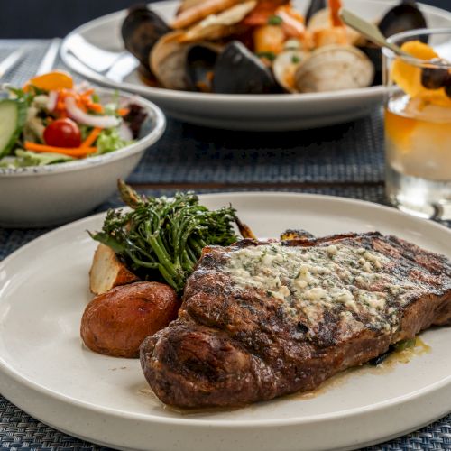 A delicious meal with a steak, potatoes, and greens, a bowl of salad, seafood, and a cocktail on a well-set dining table.