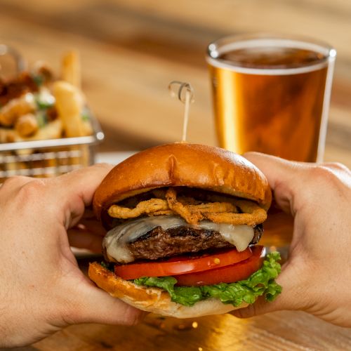 A person is holding a burger with a glass of beer and a basket of fries in the background.