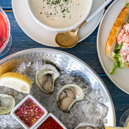 The image shows a seafood meal with oysters on ice, a bowl of chowder, a lobster roll with coleslaw, and drinks, including a red wine.