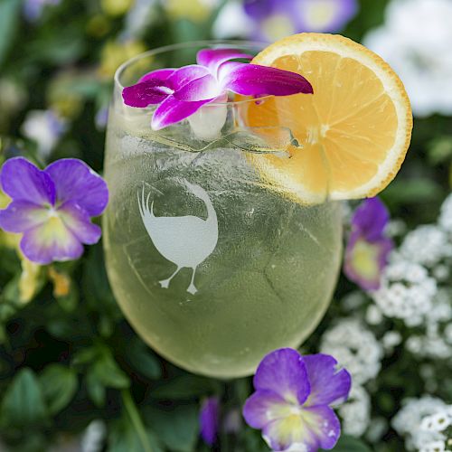 A cocktail garnished with a slice of orange and a purple flower, set against a background of lush green leaves and vibrant purple and white flowers.