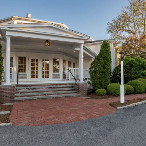 A welcoming white house with brick steps, framed by greenery, lanterns, and a curved driveway, under a clear sky and lush trees.