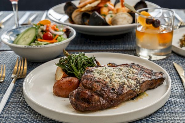 A plate with steak, potatoes, and vegetables, a seafood dish, salad, and a cocktail on a table.