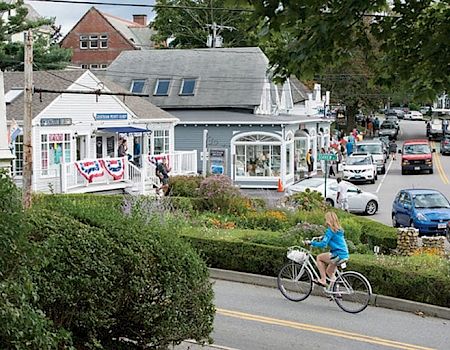 A small-town street scene with shops, a cyclist, and parked cars, surrounded by greenery and people walking on the sidewalk.