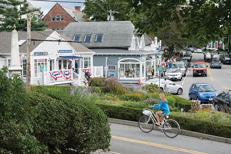 A small-town street scene with shops, a cyclist, and parked cars, surrounded by greenery and people walking on the sidewalk.