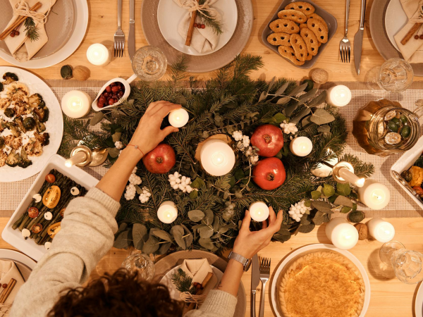 A festive table with candles, greenery, apples, and several dishes; hands reach in to place or grab items, holiday feast vibes abound.