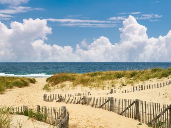 A sandy beach with wooden fences, grassy dunes, and the ocean in the background under a partly cloudy sky.
