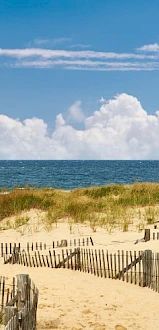 A sandy beach with wooden fences, grassy dunes, and a view of the ocean under a partly cloudy sky.