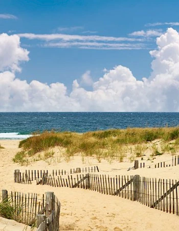 A sandy beach with wooden fences, grassy dunes, and a view of the ocean under a partly cloudy sky.