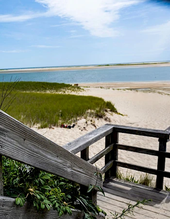 A wooden railing leading to a sandy beach and calm sea, with grassy dunes and blue sky in the background, peaceful coastal scene.