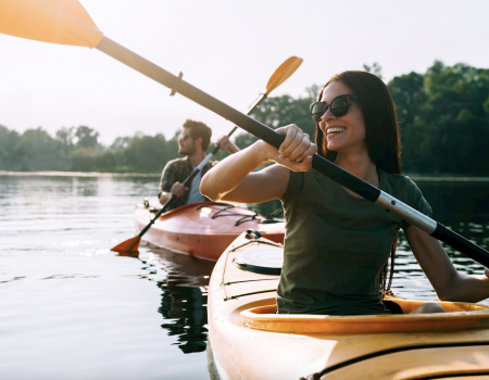 Two people kayaking on a calm lake, paddling together with orange paddles, enjoying a sunny day and scenic treeline in the background.