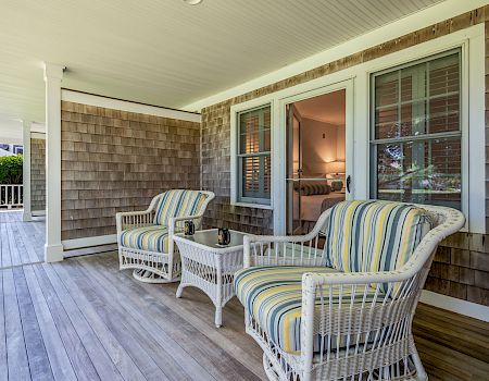 A cozy covered porch with wicker chairs, a small glass table, and striped cushions, overlooking a bright entryway through siding and windows.