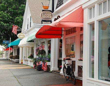 A quaint street with white storefronts and colorful awnings, hanging flower pots, a bicycle, and a sign for a manor along a sidewalk.