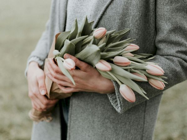 A person in a gray coat holds a bouquet of pink tulips, cradling them gently as the stems bunch together.