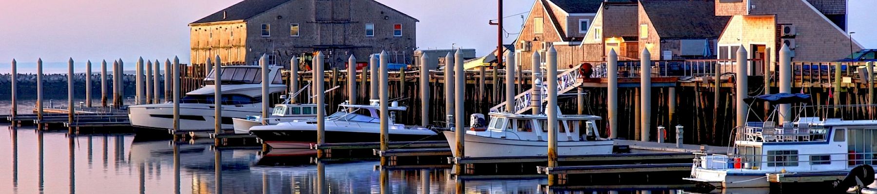 A calm harbor scene with boats docked at piers, calm water reflecting the pastel sky, and a cluster of old warehouse-like buildings along the shore.