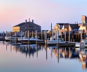 A calm harbor scene with boats docked at piers, calm water reflecting the pastel sky, and a cluster of old warehouse-like buildings along the shore.