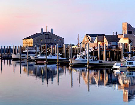 A calm harbor scene with boats docked at piers, calm water reflecting the pastel sky, and a cluster of old warehouse-like buildings along the shore.