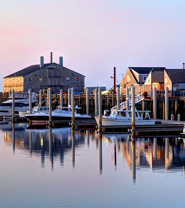 A calm harbor scene with boats docked at piers, calm water reflecting the pastel sky, and a cluster of old warehouse-like buildings along the shore.