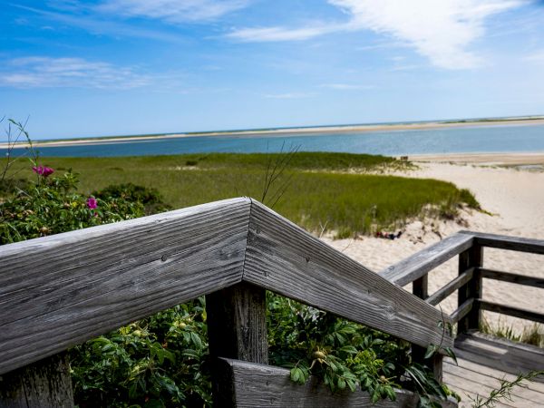 A weathered wooden railing overlooks a sandy beach with green marshes and a calm blue sea under a bright, partly cloudy sky.