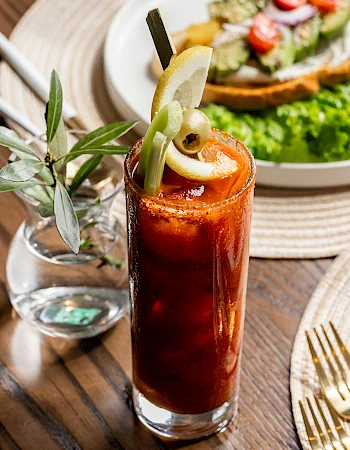 A tall Bloody Mary topped with a lime wedge, celery stick, and olive, drink in a glass, with a leafy green salad sandwich in the background.
