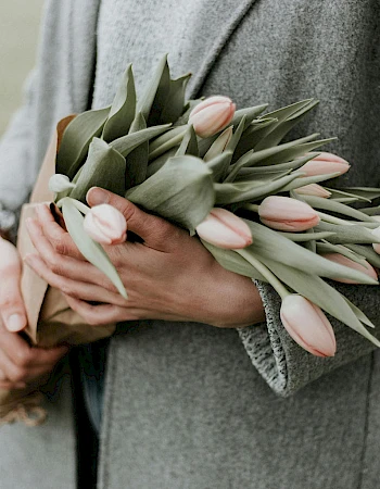 A person in a gray coat holds a bouquet of pale pink tulips, wrapped with brown paper, outdoors.