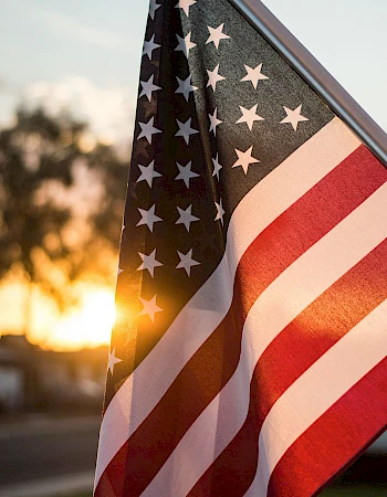 An American flag waves in the sunset, stars and stripes bright against a warm glow, patriotic and peaceful, dripping with golden light.