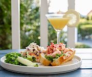 A plate with a lobster-roll sandwich, greens, and cucumber slices, plus a lemon-dusted cocktail on a sunny outdoor table.