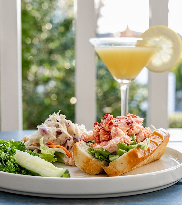 A plate with a lobster-roll sandwich, greens, and cucumber slices, plus a lemon-dusted cocktail on a sunny outdoor table.