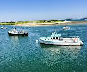 Two boats are docked in clear blue water near a sandy beach and grassy shoreline under a bright blue sky.