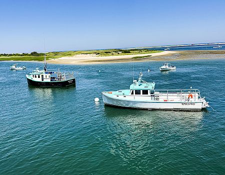 Two boats are docked in clear blue water near a sandy beach and grassy shoreline under a bright blue sky.