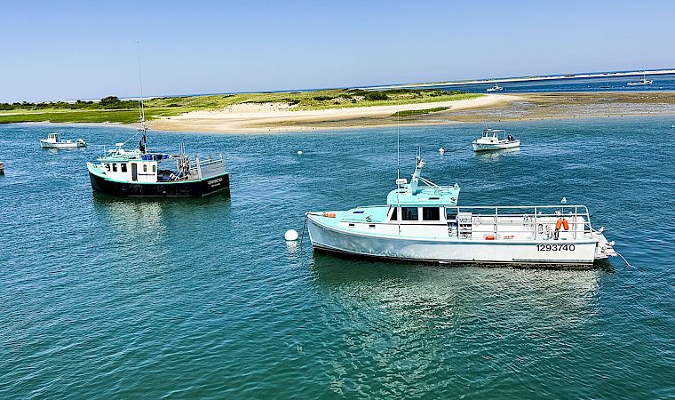 Two boats are docked in clear blue water near a sandy beach and grassy shoreline under a bright blue sky.