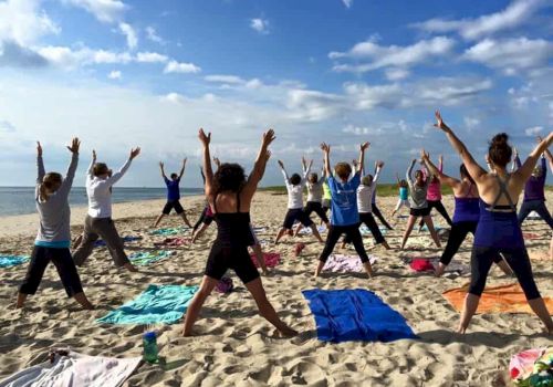 A group stretches in a yoga class on a sunny beach, arms up on colorful mats.