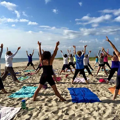 A group stretches in a yoga class on a sunny beach, arms up on colorful mats.
