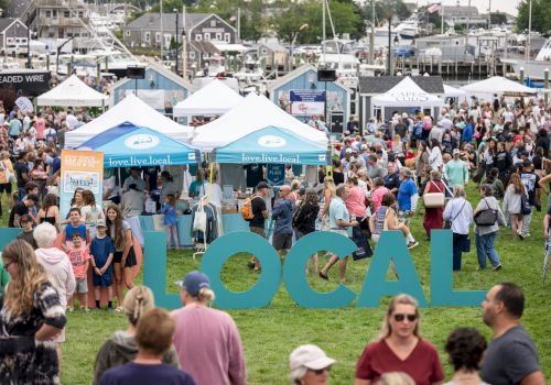 A busy outdoor &ldquo;love.live.local&rdquo; festival with lots of people in booths and tents by a marina, with big blue &ldquo;LOCAL&rdquo; letters.