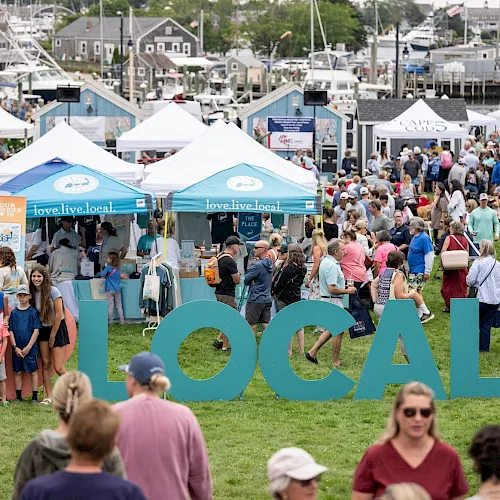 A busy outdoor &ldquo;love.live.local&rdquo; festival with lots of people in booths and tents by a marina, with big blue &ldquo;LOCAL&rdquo; letters.